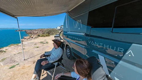 Scopri la Sardegna in van Due persone sedute vicino a un camper blu con vista sul mare