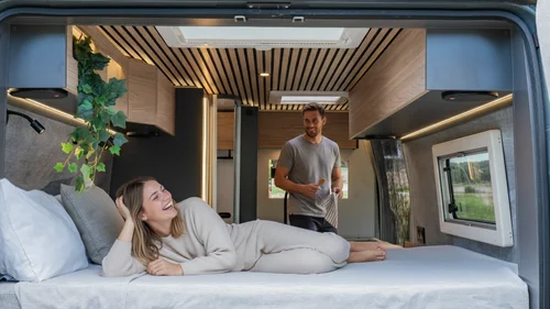 Scopri la Sardegna in van Coppia sorridente nel letto di un camper moderno e accogliente.