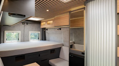 Scopri la Sardegna in van Interno moderno di un camper con letto, specchio e seduta chiara